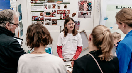 Ein Mädchen präsentiert ihr wissenschaftliches Forschungsprojekt auf dem Wettbewerb 'Jugend forscht', umgeben von mehreren Zuhörern, während sie vor einer Tafel mit Projektinformationen und Fotos auf einem Plakat steht.