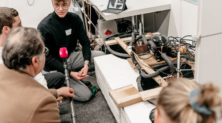 Ein junger Mann in einem schwarzen Pullover hockt mit einem Reporter und anderen Zuhörern um eine technische Apparatur, die sein Projekt auf dem Wettbewerb 'Jugend forscht' darstellt, während ein Interview mit ihm und seiner Gruppe geführt wird.
