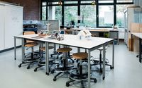 Ein heller Laborraum mit einem zentralen Arbeitstisch, umgeben von mehreren Hockern. Auf dem Tisch stehen Laptops, ein Modell eines Windrads und weitere technische Geräte.