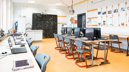 Der Computerraum des SFZs mit modernen Arbeitsplätzen, an denen Computer und Laptops aufgereiht sind, wartet auf die nächsten Schüler, die hier arbeiten werden. An der Wand im Hintergrund sind Tafelzeichnungen und Notizen zu sehen.