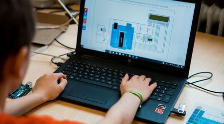 Ein Schüler arbeitet konzentriert an einem Laptop, auf dem ein Schaltplan für ein technisches Projekt angezeigt wird. Neben dem Laptop liegen eine Schaltplatine und ein USB-Stick, die Teil der technischen Ausrüstung sind, die für das Projekt verwendet wir