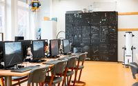 In einem modern eingerichteten Computerraum stehen mehrere Computerarbeitsplätze bereit. Im Hintergrund sind dunkle Schränke zu sehen, die mit mathematischen und naturwissenschaftlichen Formeln und Diagrammen beschrieben sind.