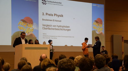 Ein Teilnehmer erhält den 3. Preis in der Kategorie 'Physik' für sein Projekt über hydrophobe Oberflächenbeschichtungen. Im Hintergrund stehen andere Teilnehmende, während das Publikum aufmerksam der Preisverleihung folgt.