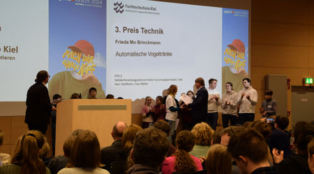 Eine Teilnehmerin nimmt auf der Bühne den 3. Preis in der Kategorie 'Technik' für ihr Projekt 'Automatische Vogeltränke' entgegen. Weitere Teilnehmende und das Publikum applaudieren während der Preisverleihung.
