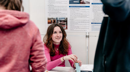 Ein Mädchen in einem pinken Pullover präsentiert ihr wissenschaftliches Projekt auf dem Wettbewerb 'Jugend forscht', während sie an einem Tisch vor einem Poster mit Projektinformationen sitzt und eines ihrer Produkte vorführt.