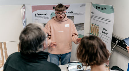 Ein Schüler in einem beigefarbenen Hoodie steht vor einem Stand mit dem Projekt 'Bee Safe' auf dem Wettbewerb 'Jugend forscht' und erklärt seine Arbeit einer Gruppe interessierter Zuhörer.