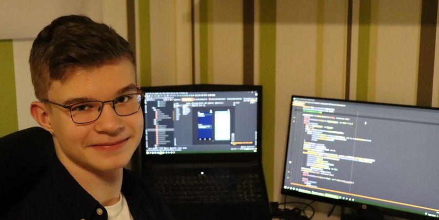 Ein junger Mann mit Brille sitzt vor zwei Bildschirmen, die Programmiercode und eine Benutzeroberfläche anzeigen. Er lächelt in die Kamera.