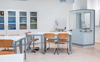 Ein heller, moderner Laborraum mit mehreren Tischen und Holzstühlen. Im Hintergrund stehen verschlossene Glasschränke mit Labormaterialien sowie ein Abzugsschrank.
