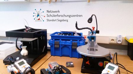 Ein Tisch mit einem 3D-Drucker, Werkzeugkasten, Bohrmaschine und zwei kleinen Robotern. Im Hintergrund hängt ein Logo mit der Aufschrift „Netzwerk Schülerforschungszentren Standort Segeberg.