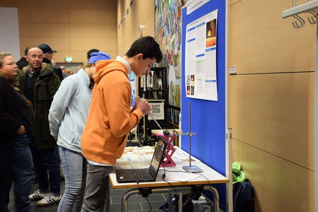 Ein junger Forscher des Schülerforschungszentrums Kieler Forschungswerkstatt führt an seinem Stand einen Versuch durch, der Teil seines Projekts ist, während interessierte Besucher aufmerksam zuschauen.