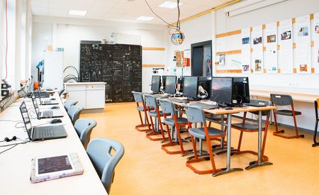 Der Computerraum des SFZs mit modernen Arbeitsplätzen, an denen Computer und Laptops aufgereiht sind, wartet auf die nächsten Schüler, die hier arbeiten werden. An der Wand im Hintergrund sind Tafelzeichnungen und Notizen zu sehen.