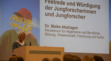 Dr. Maike Abshagen hält eine Festrede zur Würdigung der Jungforscherinnen und Jungforscher beim Wettbewerb 'Jugend forscht', während im Hintergrund eine Präsentation mit dem Titel ihrer Rede und einem dazugehörigen Bild projiziert wird.