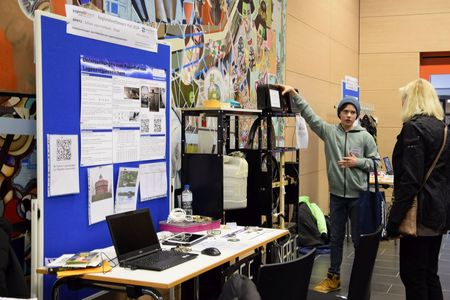Ein Teilnehmer des Schülerforschungszentrums Kieler Forschungswerkstatt erklärt einer Besucherin an seinem Stand die Funktionsweise seines Projekts, gestützt durch Poster und technische Geräte.