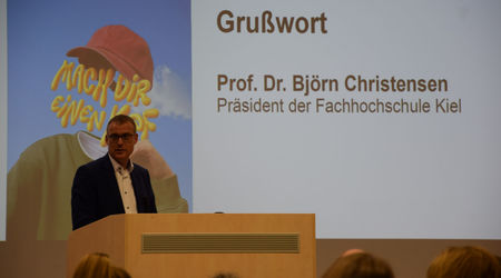 Prof. Dr. Björn Christensen, Präsident der Fachhochschule Kiel, hält ein Grußwort auf der Bühne, während im Hintergrund der Titel seiner Ansprache und ein Bild mit dem Motto 'Mach dir einen Kopf' projiziert werden.