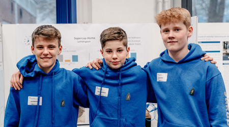 Drei Jungen in blauen Kapuzenpullovern stehen eng beieinander und lächeln in die Kamera, bei dem sie ihr gemeinsames Forschungsprojekt auf dem Wettbewerb 'Jugend forscht' präsentieren.