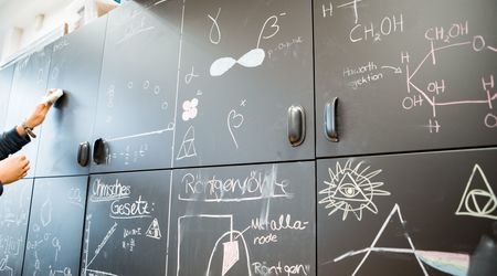 Eine Wand aus Tafeln ist mit verschiedenen wissenschaftlichen Skizzen und Formeln bedeckt, die Konzepte aus der Physik und Chemie darstellen.