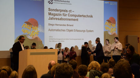 Ein Teilnehmer erhält den Sonderpreis für ein Jahresabonnement des Magazins 'c't' für sein Projekt 'Automatisches Objekt Erfassungs System' auf der Bühne. Andere Teilnehmende applaudieren im Hintergrund, während das Publikum die Szene beobachtet.