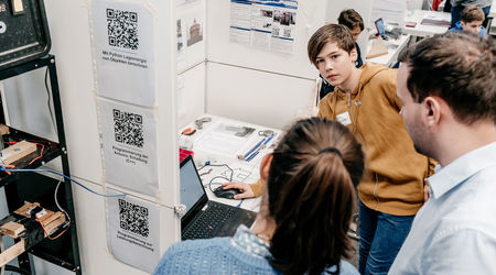 Ein Junge in einem braunen Kapuzenpullover steht neben einem Laptop und mehreren Projektplakaten, während er von mehreren Zuhörern umgeben ist.