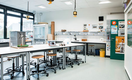 Ein heller, geräumiger Laborraum mit mehreren Arbeitstischen und Hockern. Auf den Tischen befinden sich ein Laptop, ein Modell und verschiedene wissenschaftliche Geräte. An den Wänden hängen Steckdosen und Gasanschlüsse.
