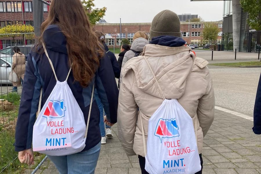 Zwei Personen von hinten gesehen, die mit weißen Turnbeuteln mit der Aufschrift 'Volle Ladung... MINT Akademie' auf dem Rücken an einem Gebäude entlang gehen.
