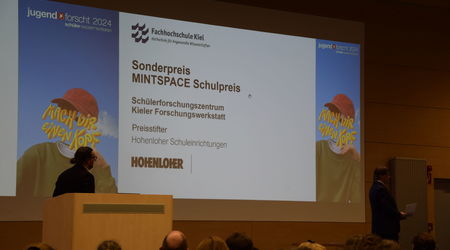 Der Sonderpreis MINTSPACE Schulpreis wird an das Schülerforschungszentrum Kieler Forschungswerkstatt überreicht, während der Redner den Preis auf der Bühne präsentiert und das Publikum die Ehrung verfolgt.