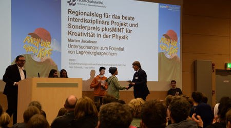 Ein Teilnehmer erhält auf der Bühne den Regionalsieg für das beste interdisziplinäre Projekt und den Sonderpreis plusMINT für Kreativität in der Physik. Im Hintergrund applaudieren weitere Teilnehmende, während das Publikum die Auszeichnung verfolgt.