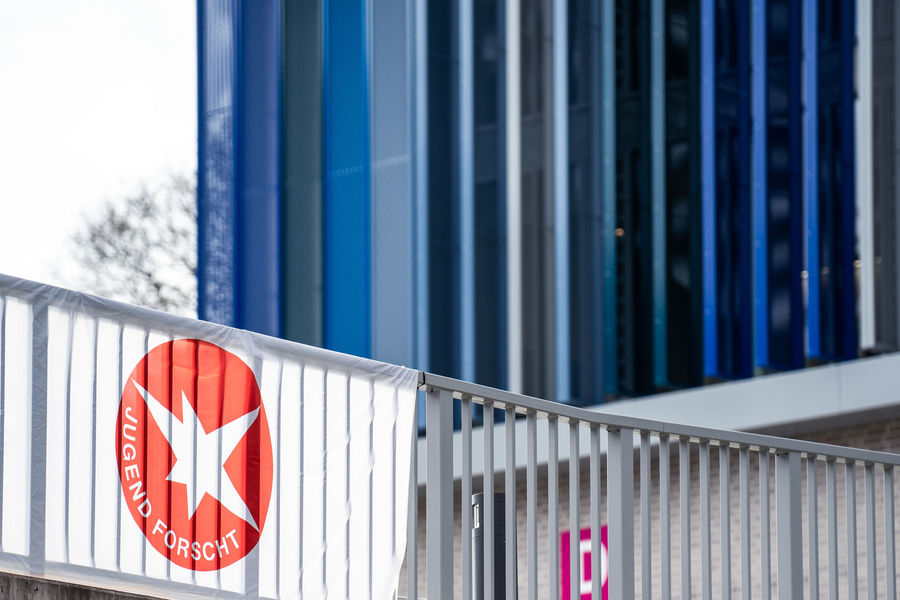 Blick auf ein Gebäude mit blauen Fassadenstreifen, im Vordergrund ein Geländer mit einem rot-weißen 'Jugend forscht'-Banner.