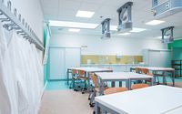 Ein heller, moderner Laborraum mit mehreren Arbeitstischen und Holzstühlen. An der linken Wand hängen weiße Laborkittel an Haken. An der Rückwand befinden sich Arbeitsflächen mit Waschbecken und Schränken.
