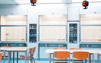 In diesem Laborraum stehen mehrere Tische mit Holzstühlen vor einer Reihe von großen Abzugsschränken, die an der Rückwand installiert sind. An der Decke sind rote Anschlüsse für Laborgeräte montiert.