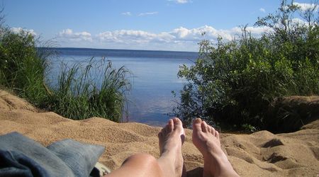 Person liegt am sandigen Ufer eines Sees mit Blick auf das ruhige Wasser und den blauen Himmel, umgeben von Büschen und Gräsern.