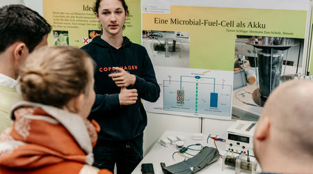 Ein Jugendlicher in einem dunkelblauen Hoodie steht vor seinem Projektstand mit Poster zum Thema 'Microbial-Fuel-Cell als Akku' und erklärt sein Projekt einer Gruppe interessierter Zuhörer auf dem Wettbewerb 'Jugend forscht'.