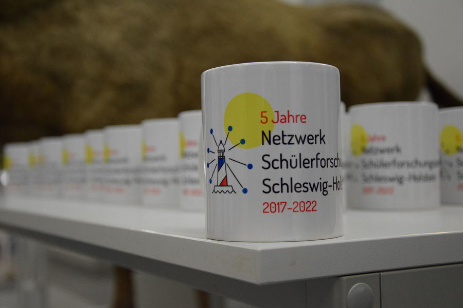 Eine Reihe von Tassen, die das 5-jährige Jubiläum des Netzwerks Schülerforschungszentren Schleswig-Holstein (2017–2022) feiern, stehen auf einem Tisch bereit, jede mit einem symbolischen Design eines Leuchtturms und der Aufschrift.