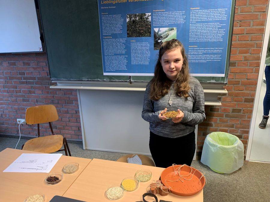 Ein Mädchen steht vor einer Präsentationstafel in einem Klassenraum und hält eine Schale mit Körnern in den Händen. Auf dem Tisch vor ihr liegen mehrere Schalen mit verschiedenen Getreidesorten und ein oranger Topfuntersetzer.