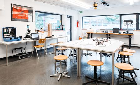 Ein heller, moderner Laborraum mit mehreren Arbeitstischen und Hockern. Auf den Tischen liegen Roboter und technische Geräte. Im Hintergrund stehen weitere Arbeitsplätze mit Werkzeugen, Computern und Geräten. Große Fenster lassen viel Tageslicht herein.
