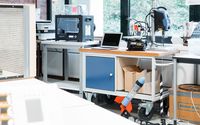 Ein Labortisch mit einem Laptop, einem 3D-Drucker und weiteren technischen Geräten. Unter dem Tisch steht eine kleine Modellrakete. Im Hintergrund sind große Fenster, die viel Tageslicht hereinlassen, sowie weitere Laborgeräte und Materialien zu sehen.