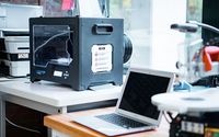 Ein Nahaufnahme eines Labortischs mit einem 3D-Drucker und einem offenen Laptop. Neben dem Laptop befindet sich ein weiteres technisches Gerät.