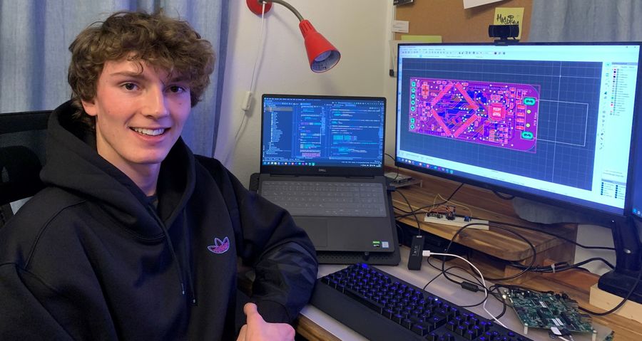 Ein junger Mann sitzt lächelnd an einem Schreibtisch mit zwei Bildschirmen. Auf einem Bildschirm ist ein Schaltplan-Layout zu sehen, auf dem anderen Bildschirm sind Programmiercodes angezeigt.
