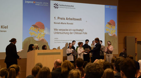 Die Gewinnerin des 1. Platzes in der Kategorie 'Arbeitswelt' nimmt ihren Preis für das Projekt 'Wie verpacke ich nachhaltig? Untersuchung der Chitosan Folie' auf der Bühne entgegen, während andere Teilnehmende der Kategorie im Hintergrund stehen und das P