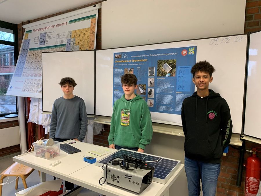Drei Jugendliche stehen vor einer Präsentationstafel in einem Klassenraum. Auf dem Tisch vor ihnen befinden sich Solarmodule und technische Geräte.