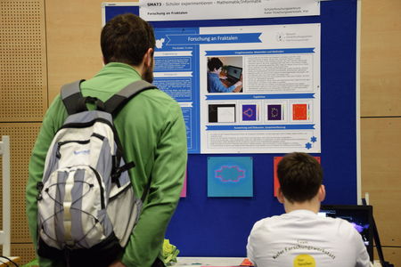 Ein Teilnehmer des Schülerforschungszentrums Kieler Forschungswerkstatt erklärt einem interessierten Besucher sein Forschungsprojekt über Fraktale, unterstützt von einem detaillierten Poster und Präsentationsmaterialien.