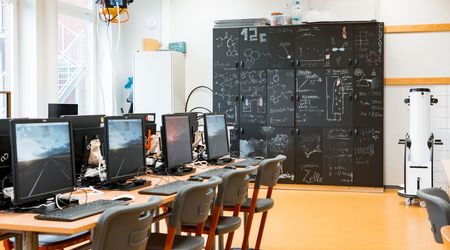 In einem modern eingerichteten Computerraum stehen mehrere Computerarbeitsplätze bereit. Im Hintergrund sind dunkle Schränke zu sehen, die mit mathematischen und naturwissenschaftlichen Formeln und Diagrammen beschrieben sind.