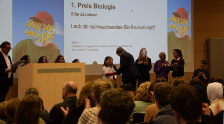 Die Gewinnerin des 1. Platzes in der Kategorie 'Biologie' nimmt ihren Preis für das Projekt 'Laub als nachwachsendes Bio-Baumaterial?!' auf der Bühne entgegen, während andere Teilnehmende applaudieren und das Publikum die Auszeichnung verfolgt.