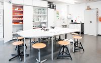 Ein heller Laborraum mit einem großen, zentralen Arbeitstisch, auf dem mehrere kleine Roboter stehen. Um den Tisch sind Hocker verteilt. Im Hintergrund befinden sich offene Schränke mit Werkzeugen und Materialien.