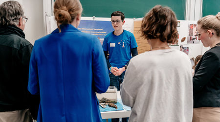 Ein Junge in einem blauen T-Shirt präsentiert sein Forschungsprojekt, umgeben von vier Zuhörern, während er vor einer Tafel mit Projektpostern steht.