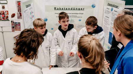Eine Gruppe von drei Jungen in weißen Laborkitteln präsentiert ihr Forschungsprojekt mit dem Titel 'Raketendünger' auf dem Wettbewerb 'Jugend forscht', umgeben von mehreren Zuhörern vor ihren Projektpostern.