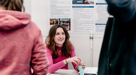 Ein Mädchen in einem pinken Pullover präsentiert ihr wissenschaftliches Projekt auf dem Wettbewerb 'Jugend forscht', während sie an einem Tisch vor einem Poster mit Projektinformationen sitzt und eines ihrer Produkte vorführt.