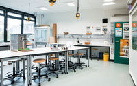 Ein heller, geräumiger Laborraum mit mehreren Arbeitstischen und Hockern. Auf den Tischen befinden sich ein Laptop, ein Modell und verschiedene wissenschaftliche Geräte. An den Wänden hängen Steckdosen und Gasanschlüsse.