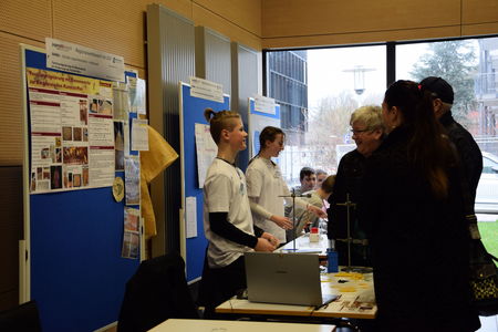 Ein junger Forscher des Schülerforschungszentrums Kieler Forschungswerkstatt diskutiert sein Projekt mit Besuchern an seinem Stand, während er die Details seiner Forschung und die dazugehörigen Materialien präsentiert.