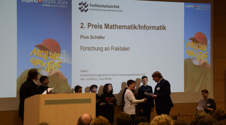 Der 2. Preis in der Kategorie 'Mathematik/Informatik' wird an einen Teilnehmer für sein Projekt zur 'Forschung an Fraktalen' überreicht. Im Hintergrund stehen weitere Teilnehmende der Kategorie, während das Publikum die Auszeichnung verfolgt.