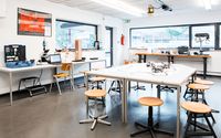 Ein heller, moderner Laborraum mit mehreren Arbeitstischen und Hockern. Auf den Tischen liegen Roboter und technische Geräte. Im Hintergrund stehen weitere Arbeitsplätze mit Werkzeugen, Computern und Geräten. Große Fenster lassen viel Tageslicht herein.